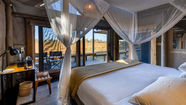 Double bed in a safari tent with mosquito netting, a desk and chair leading to open doors and a deck overlooking the desert