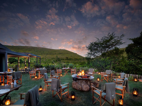 Chairs and tables laid out on a deck at sunset with green hills in the back ground and cloudy sky with lanterns all around