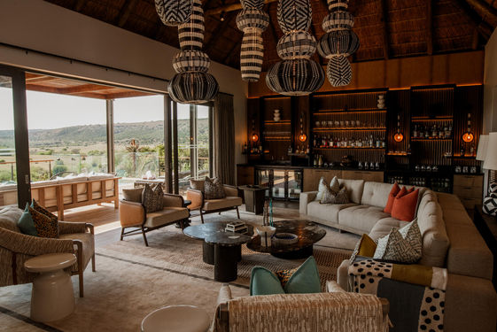 Large Drawing room with curved sofa and armchairs under hanging lights and a wooden roof looking through glass windows over Shamwari