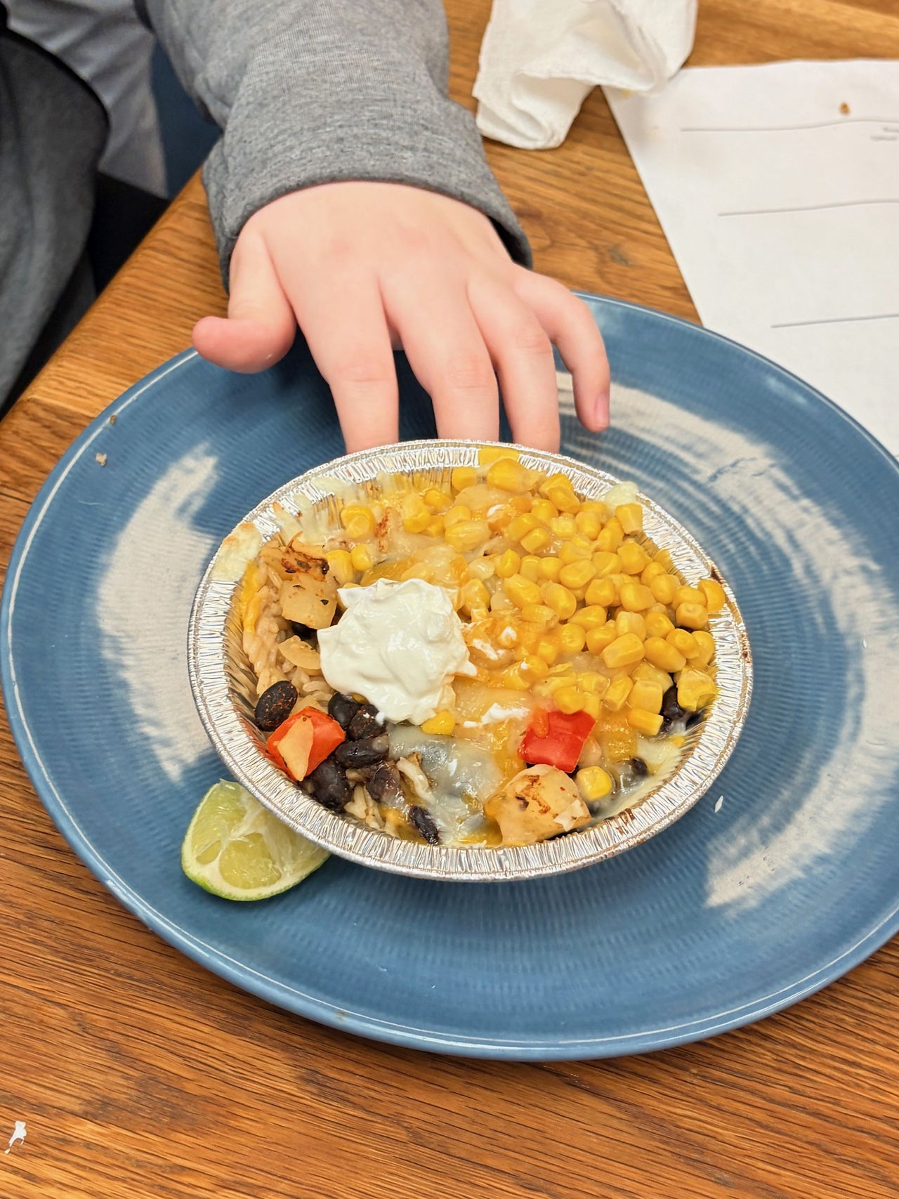 A close up photo of a freshly made burrito bowl in a small disposal foil bowl. The close-up highlight the melty cheese, corn, black beans, and sour cream topping. All on a blue plate with a small lime slice on the side. 
