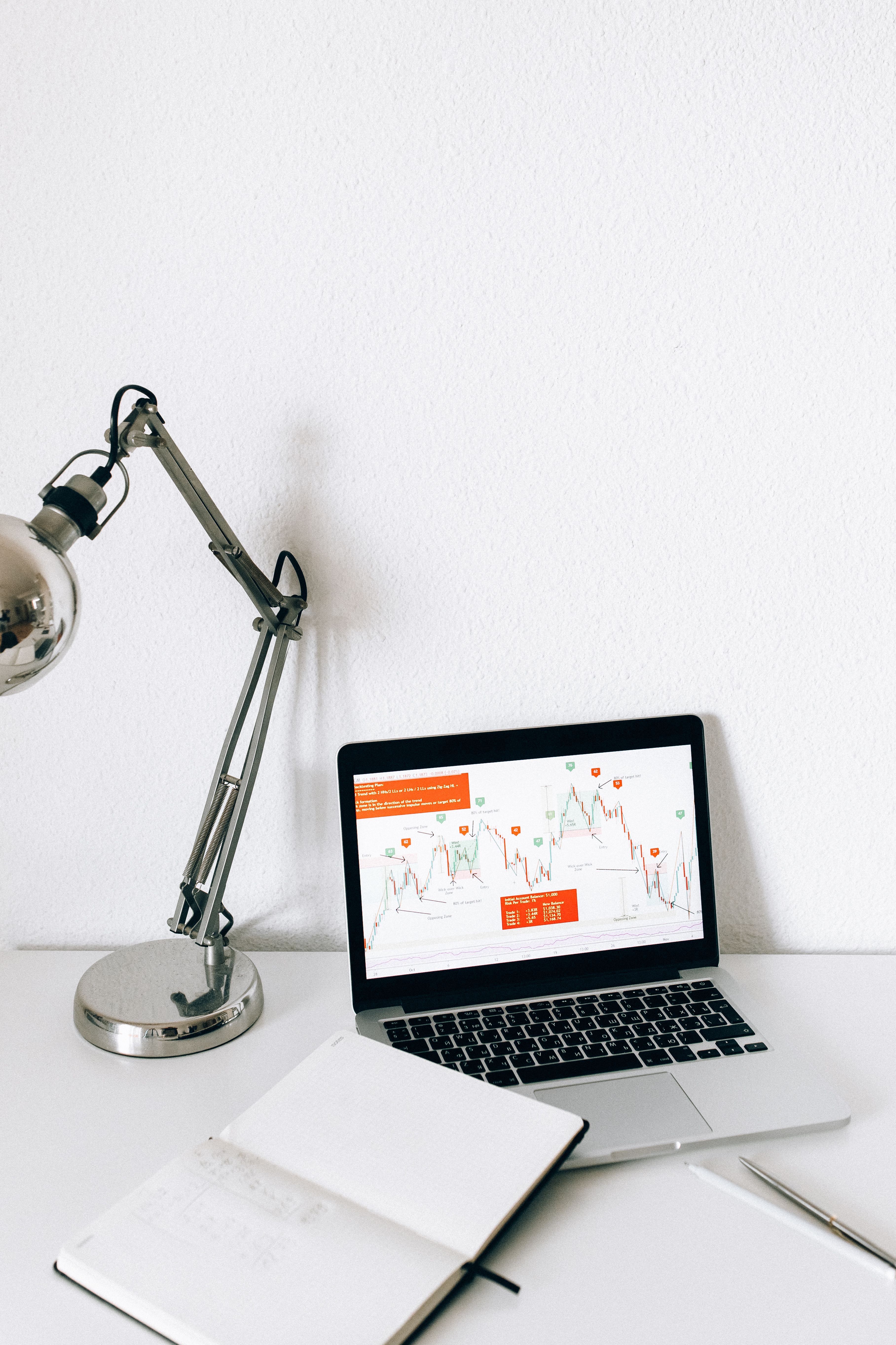 An open notebook and a desk lamp beside a laptop displaying charts.