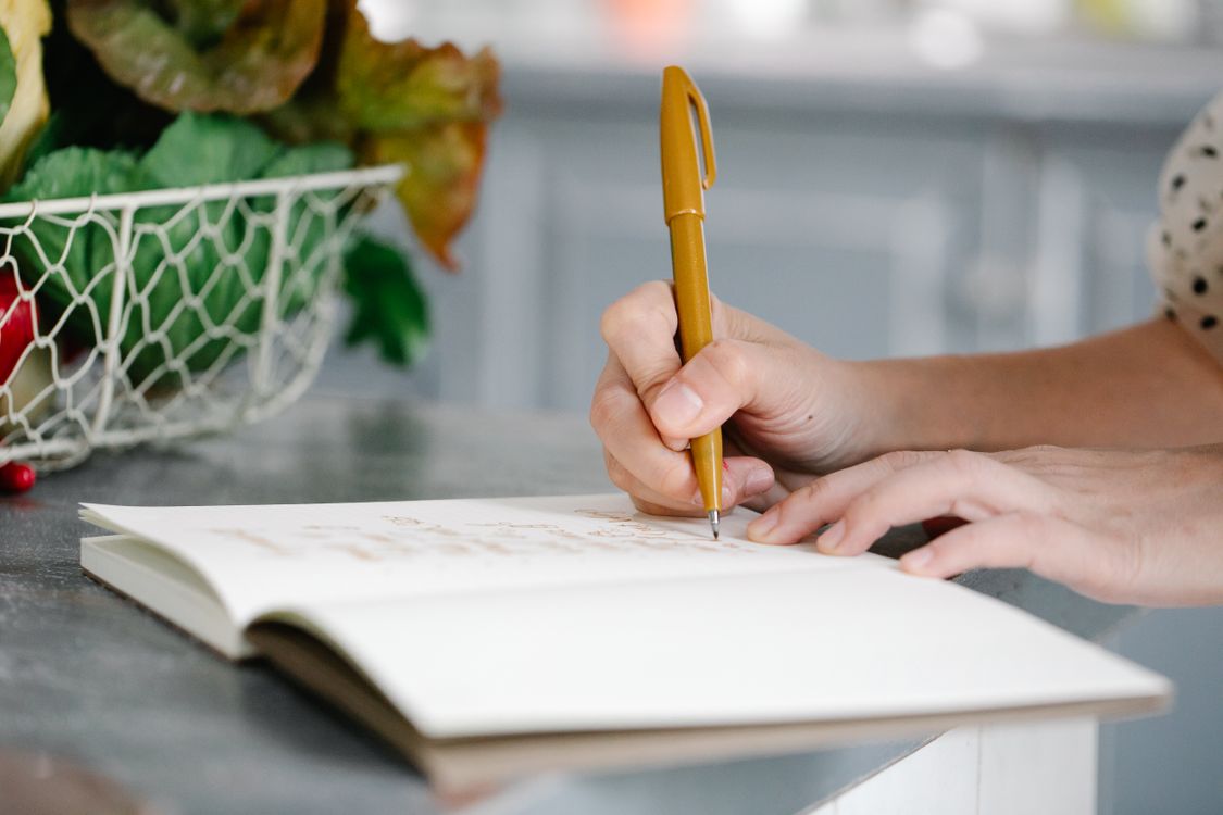 Hands writing in a notebook beside fresh produce on a kitchen counter.