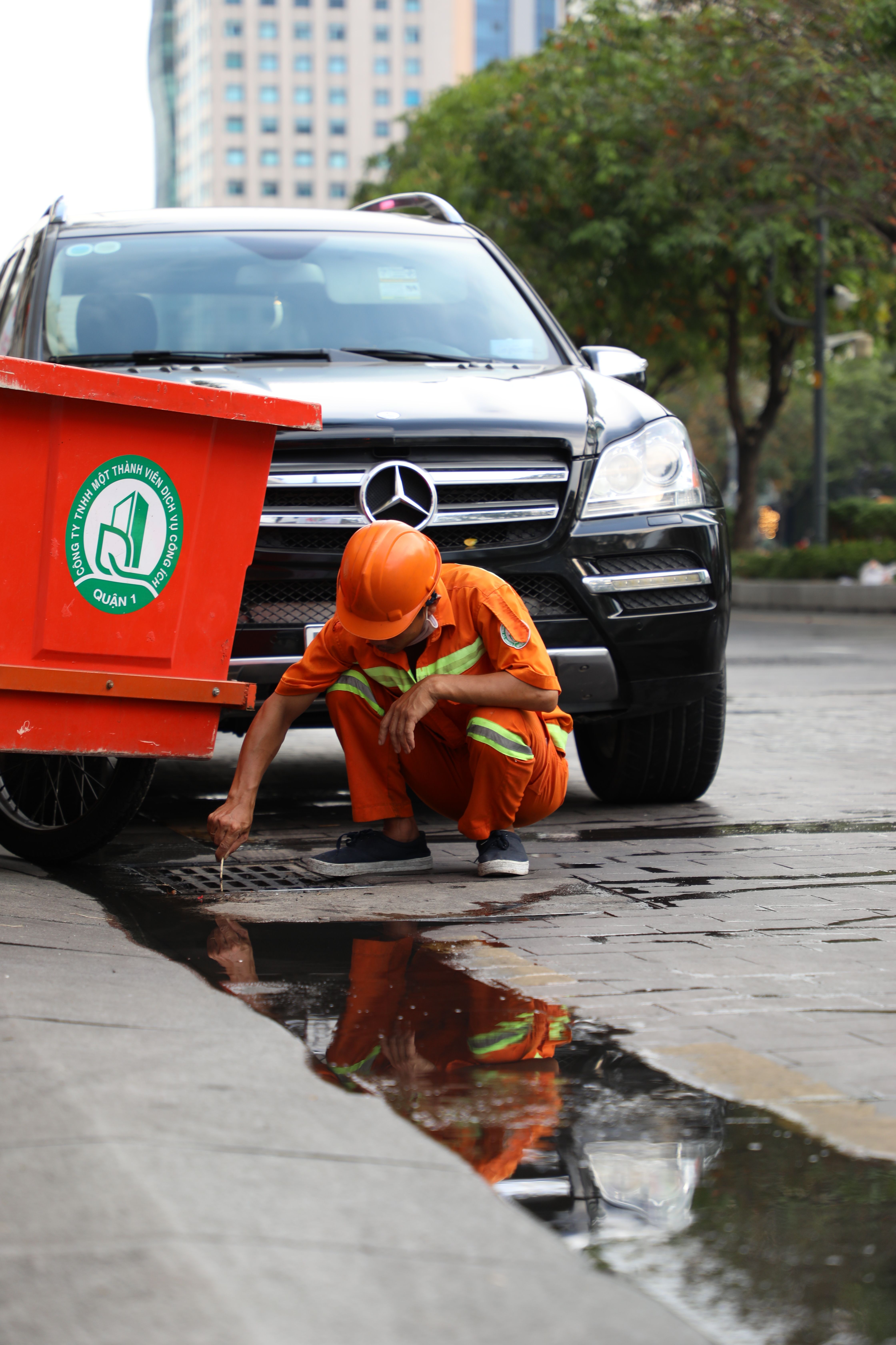 A person in orange workwear crouching beside a street drain and parked vehicle.