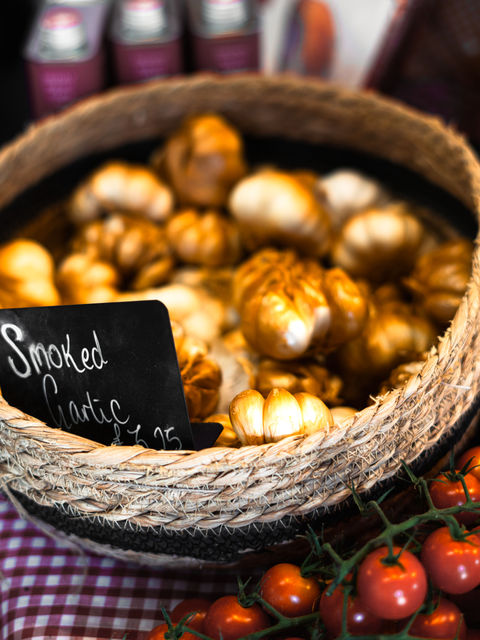 A small woven basket filled with smoked garlic, with a price tag displayed on a red chequered tablecloth.