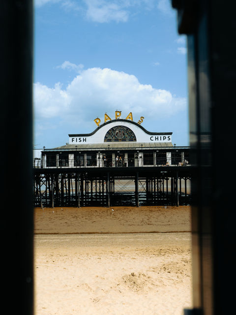 The image shows the Papas Fish & Chips shop on the Cleethorpes Pier framed between two black metal posts, with a section of sandy beach in the foreground.
