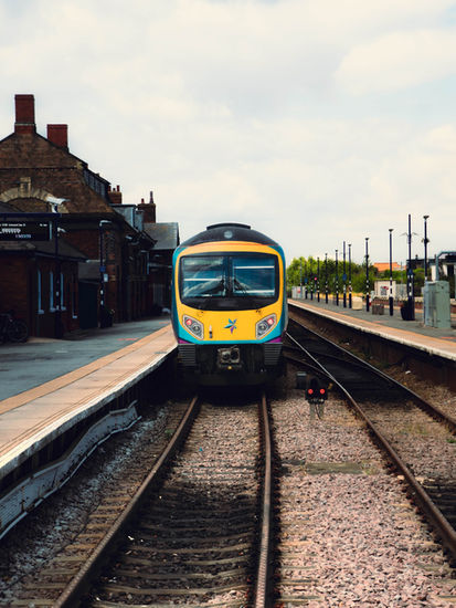This image shows a class 185 desiro diesel train from a head on perspective as it stops at a station. Surrounding the train are two sides of the station, with the left featuring a station house, and the right, a row of lamp posts and trees.