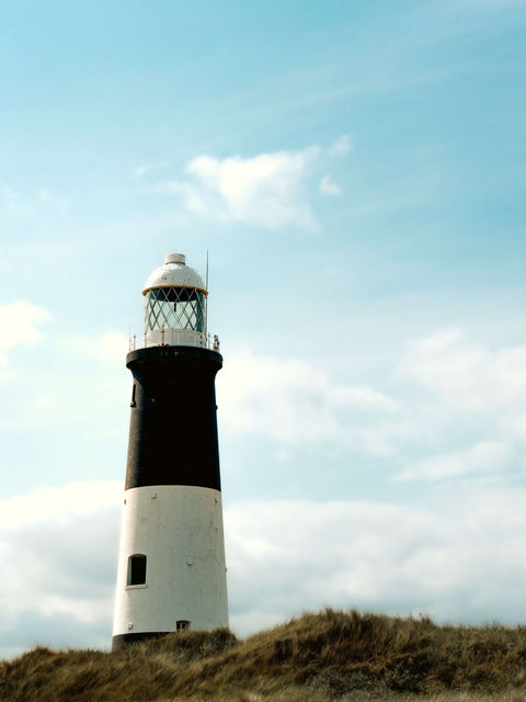 The image shows the spurn point lighthouse positioned atop a grassy dune to the left of the frame. A beautiful blue sky and light cloud coverage sits in the background.