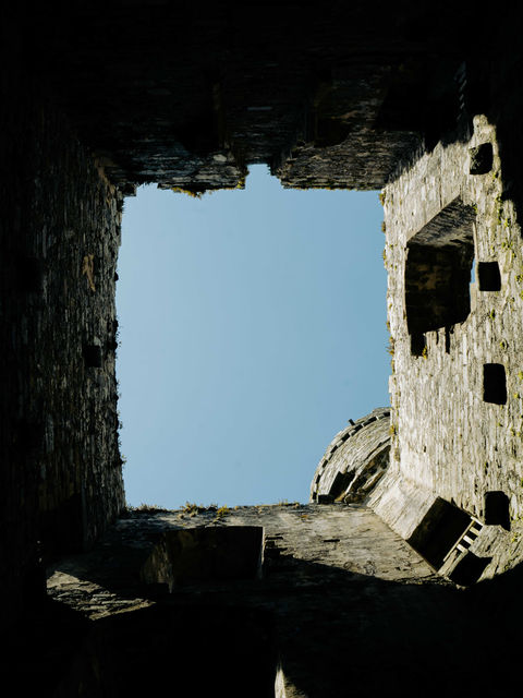 The picture depicts the inside of Castle Harlech, looking up to a blue sky with the ramparts and a tower in view.