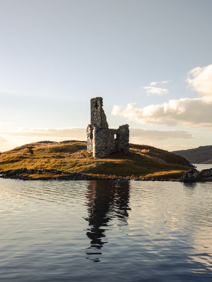 Standing alone on a rocky promontory in Loch Assynt, the ruinous Ardvreck Castle is decorated with wild plant life and surrounded by a picturesque landscape.