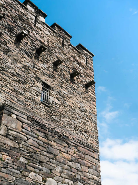 The image shows a side view of Dolwyddelan Castle from ground level, with a bright blue sky and thin cloud above.