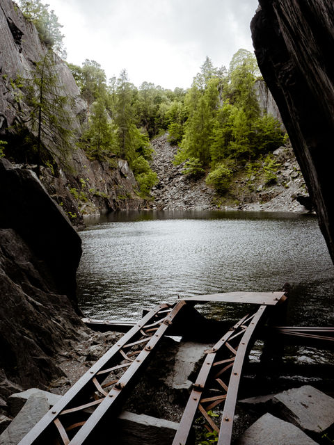 The image shows, in its foreground, a derelict piece of mining equipment sticking out over a body of water. In the background, a bank of slate with trees growing randomly among them.
