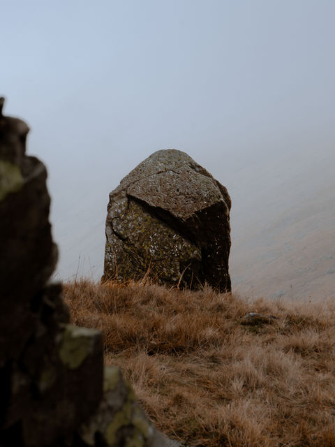 The image shows a stone resembling a church steeple, The Kirkstone, as it sits near the summit of Kirkstone pass, with a foggy backdrop.