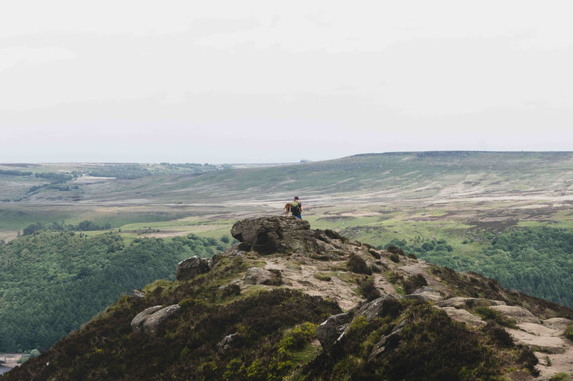 The image shows a shot of a hiker and her dog sat atop Win Hill in the Peak District as they overlook the expansive landscape of Bamford Moor.