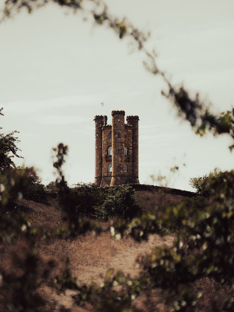 The images shows Broadway Tower from a distance between branches from a bush.