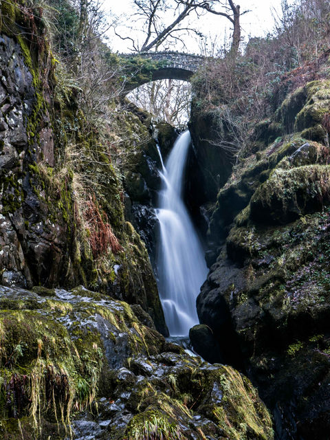 The picture is of Aira Force shot with a long exposure to give the water a smooth, silky appearance. It flows under an arched bridge with moss-covered rocks in the foreground and trees in the background.