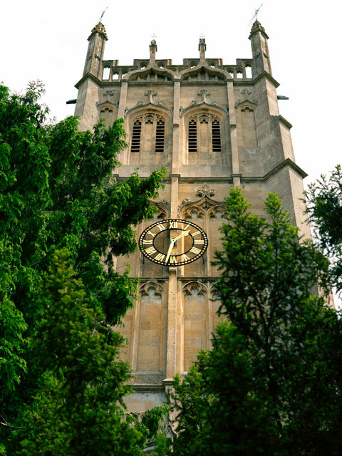 The image shows the towering Church of St James in Chipping Campden, it's clock face a striking golden colour sitting between two trees.