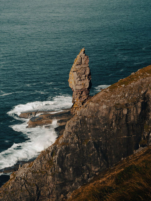 The image shows the Old Man of Stoer from a raised position, looking down onto the rock formation with waves lapping against the base of the sea stack.