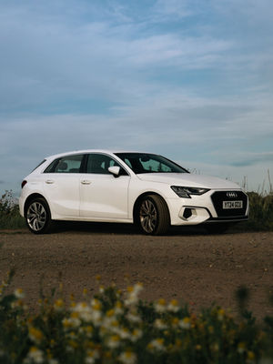 A white Audi A3 on a dirt track, with the camera capturing its right side. In the foreground, soft-focus daisies blur gently into view.