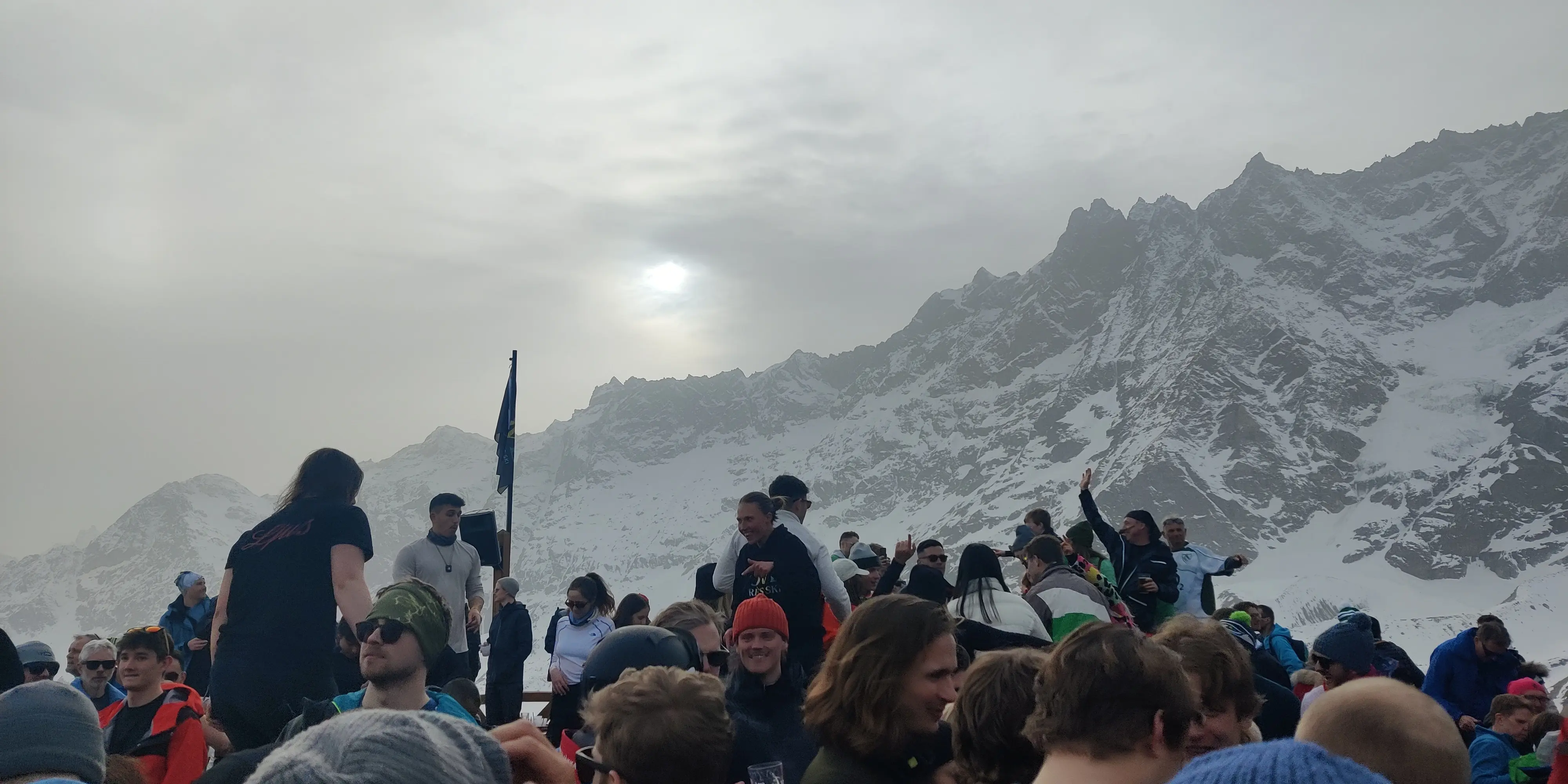 Gezellige après-ski in de Italiaanse Alpen, waar wintersporters samen feesten op een zonnig bergterras met besneeuwde bergtoppen op de achtergrond.