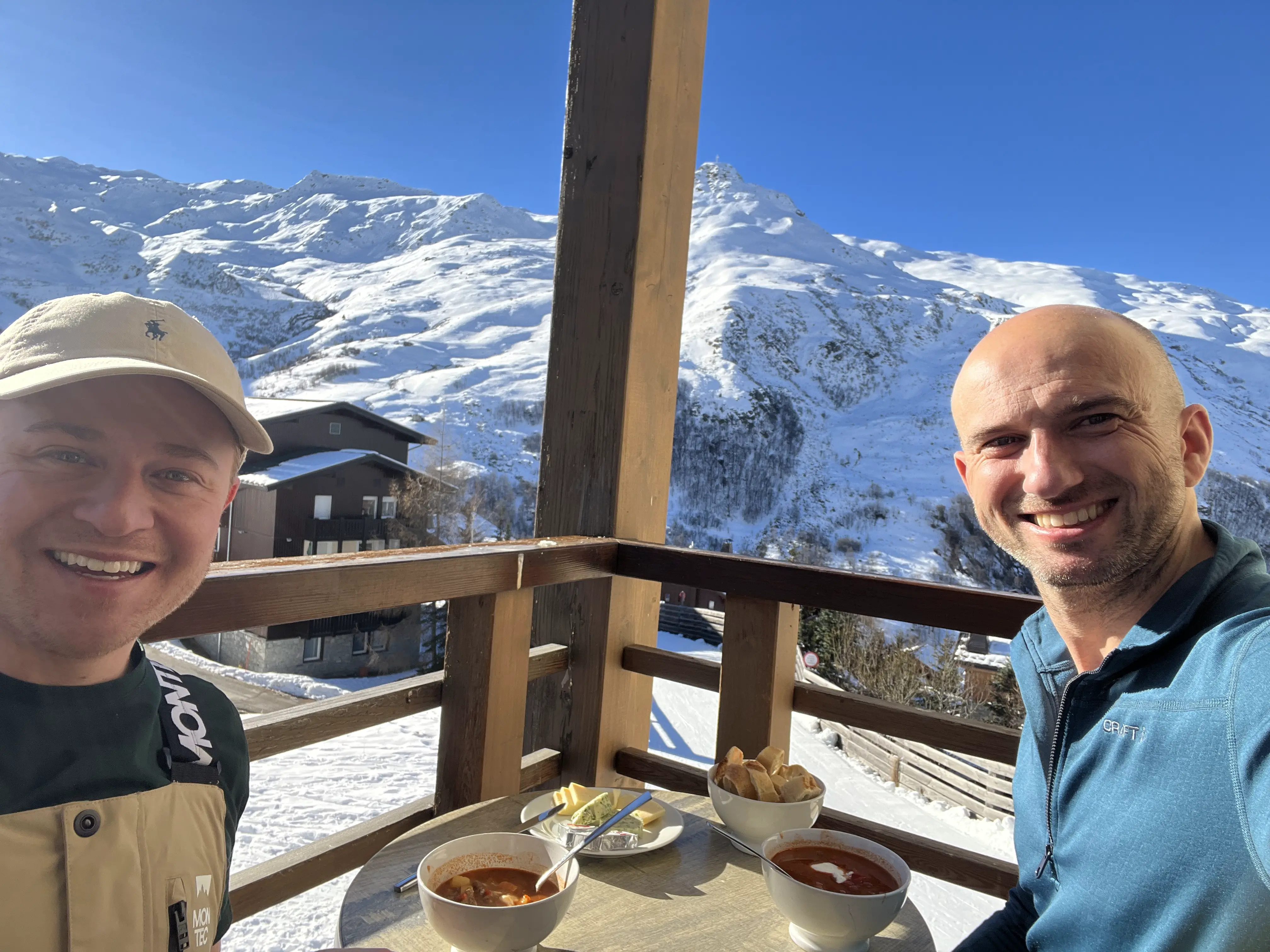 Twee mannen (de schrijver van het artikel) genietend van goulash soep op het balkon met op de achtergrond het skidorp en bergen