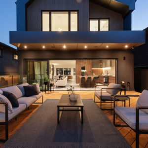 Rear patio deck with outdoor lounge seating and dining under a canopy, facing the house’s large sliding doors and lit interior.