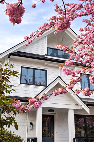 Close up of the duplex façade showing shingle cladding, dark window trim, and gable roof detailing framed by cherry blossoms.