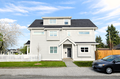 West elevation view of the duplex showing the gable roof profile, grouped windows for daylight, and light-toned siding with shingle accents along the side street.