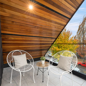 Covered upper balcony under a wood soffit with seating and glass railing, looking toward the street and treetops beneath the main gable roof.