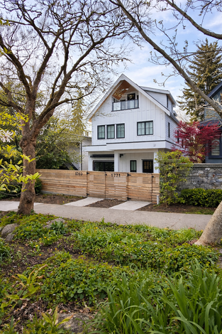 Distant curbside view of the duplex in its streetscape, showing the gable roof profile, covered entry, and landscaped front yard along the sidewalk.