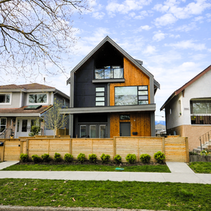 Streetscape view of the duplex showing the gable roof profile, warm wood accents, and a landscaped front yard behind low fencing.