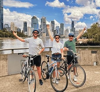 Three people each with one hand in the air.  They are holding bicycles and wearing helmets.  The city is in the background.