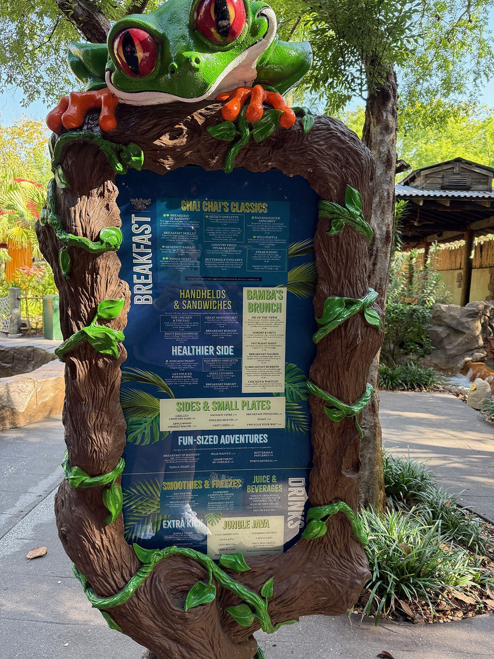 Menu board with green frog design, featuring breakfast items and sandwiches. Set in a leafy outdoor area with sunlight filtering through. This is Rainforest Cafe menu at Disney's Animal Kingdom Theme Park.