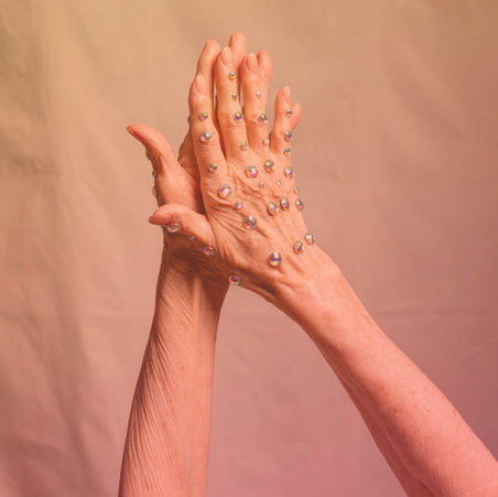 Elderly hands with colorful gems raised in prayer against a warm gradient background