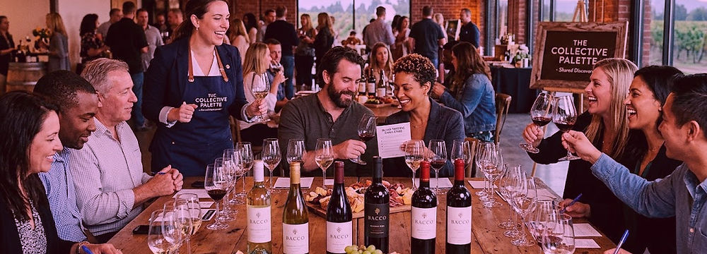 A large group enjoying a lively, interactive private wine tasting around a long rustic table in a beautifully lit event space. BACCO