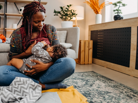 A parent sits cross-legged on a rug in a cozy living room, breastfeeding a toddler. The parent looks down gently at the child, surrounded by soft blankets.