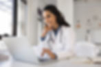 Smiling female Latin doctor wearing white medical uniform sit at desk using modern laptop,