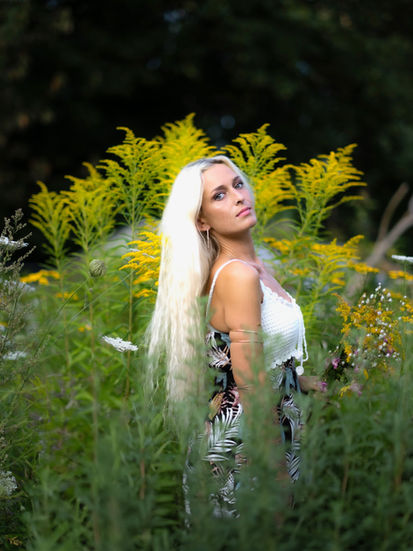 Porträt von Denise im Profil mit Blick über die Schulter vor gelbem Blütenbusch, gebräunter Rücken, blondes Haar im Sonnenlicht.