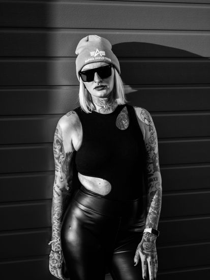 Selbstbewusstes Schwarz-Weiß-Portrait von Natalie mit Sonnenbrille, an Garagentor gelehnt, direkter Blick in die Kamera, starke Ausstrahlung