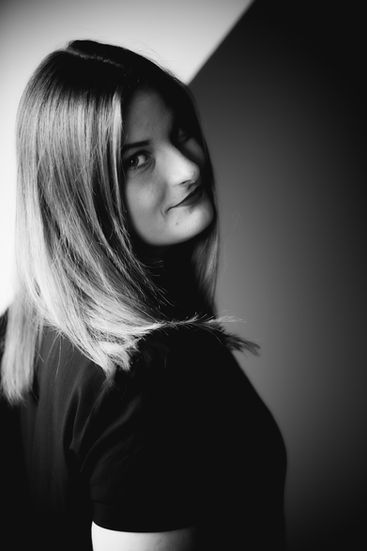 Schwarz-Weiß-Portrait einer jungen Frau im Fensterlicht, mit Schulterblick zur Kamera, aufgenommen im Dachzimmer.
