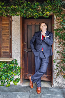 Man in a navy suit with a red tie leans thoughtfully against a wooden door, surrounded by green ivy on a stone wall. Brown shoes highlight the look.