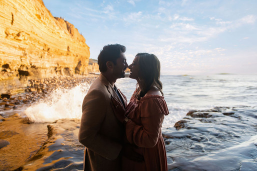 Couple embraces near a rocky shoreline at sunset, waves crashing behind them. Golden cliffs in the background, creating a romantic mood.