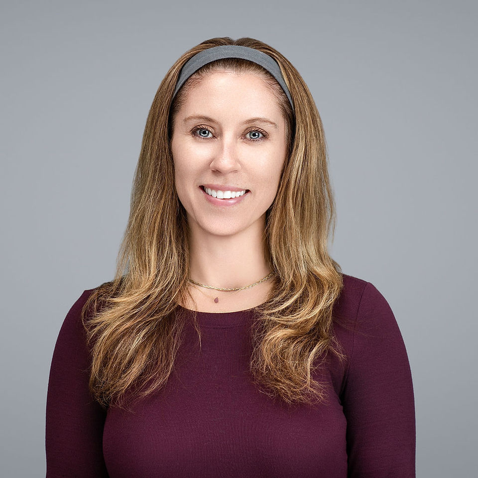 Woman with long light brown hair wearing maroon long-sleeve top and gray headband, standing against gray background, smiling.