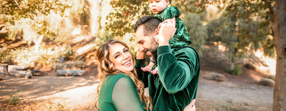 A smiling family in matching green velvet outfits; a woman, a man carrying a toddler on his shoulders, in a sunlit wooded setting.