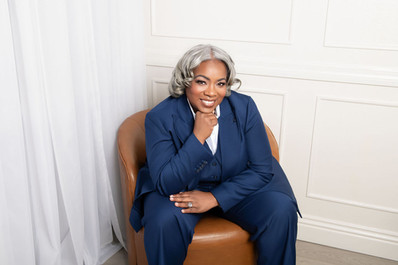 Smiling person in a blue suit sits on a brown chair, with white paneled walls and curtains in the background, exuding confidence.