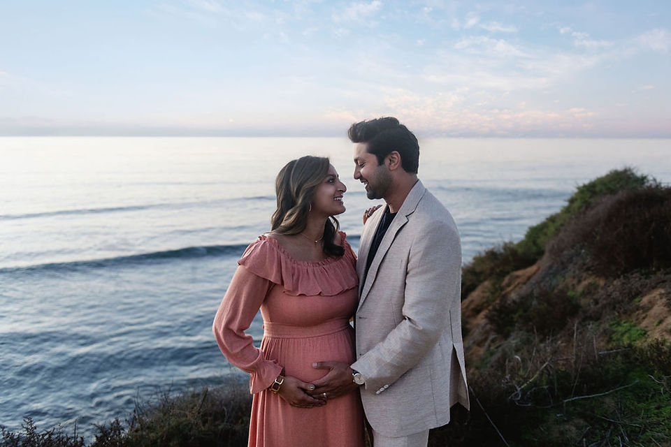 A couple by the seaside, dressed in pastel. They lovingly touch the woman's baby bump, with the ocean and sunset sky in the background.