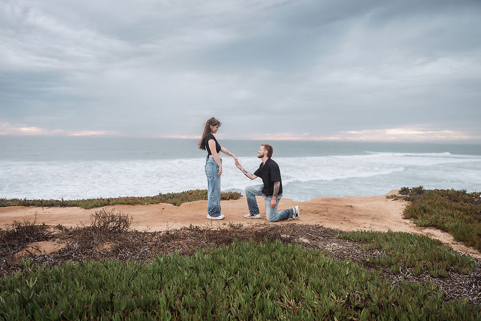 Man kneels and proposes to a woman on a coastal cliff, holding her hand under a cloudy sky at Tamarack State Beach in Carlsbad.
