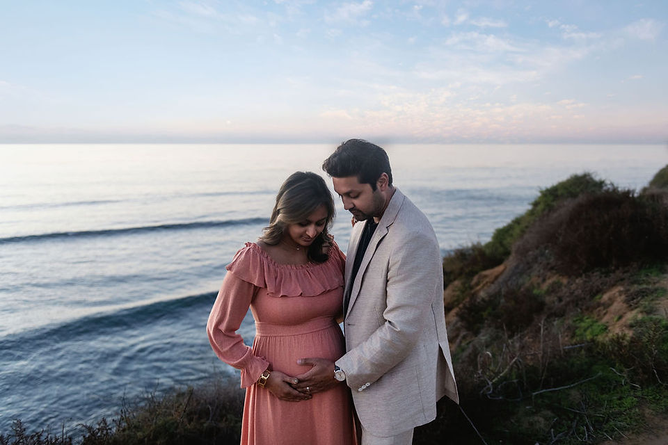 A couple embraces on a coastal cliff with a serene ocean backdrop. The woman in a pink dress holds her belly, creating a joyful, loving mood.