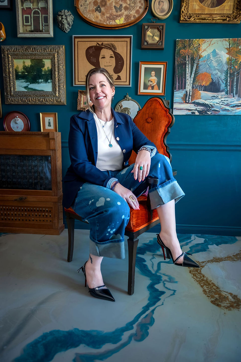 Confident female CEO in navy blazer and black heels sitting on orange chair with teal gallery wall in Temecula