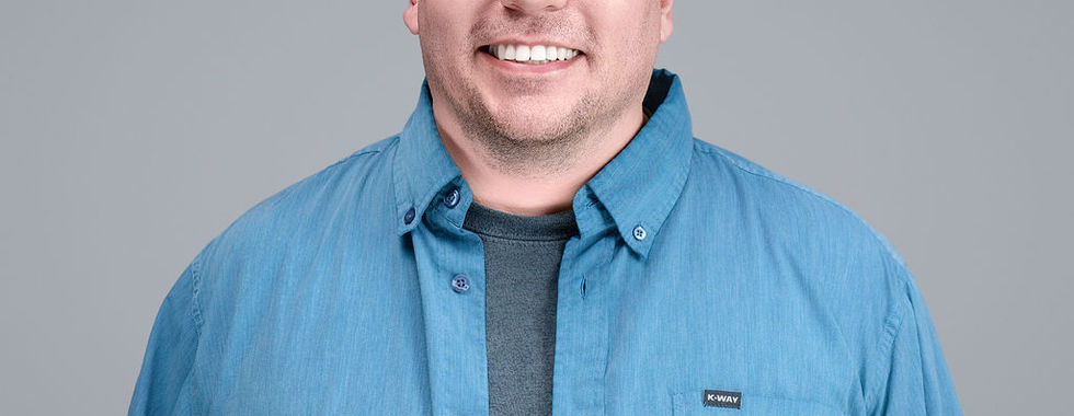 Man with short dark hair wearing light blue button-up shirt, standing against gray background, smiling