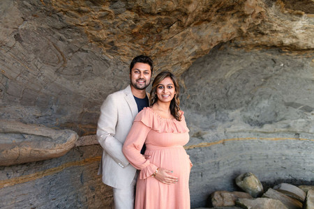 A couple poses in a rocky cave setting. The woman in a peach dress cradles her belly, while the man smiles beside her, wearing a beige suit.